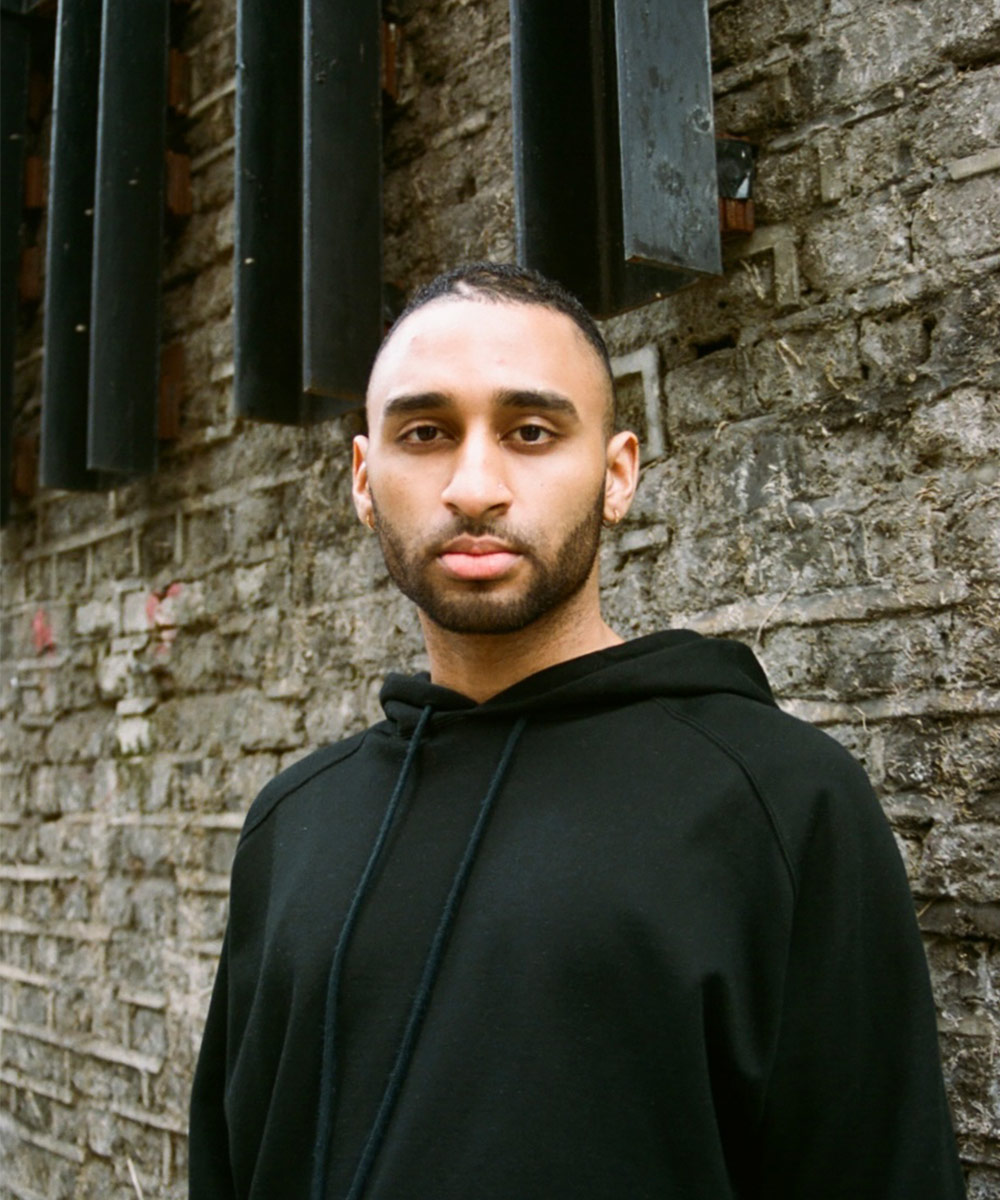 Seb Odyssey stares at the camera, in a black hoodie, against a brick wall