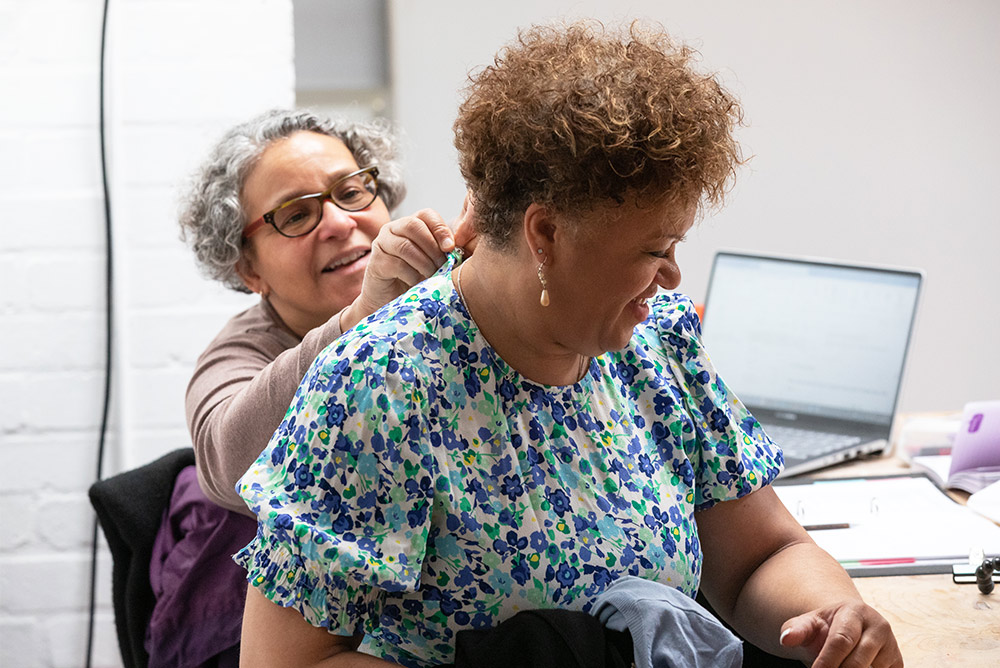 An older black-brown woman adjusts the back of a costume for MRS, an older black woman