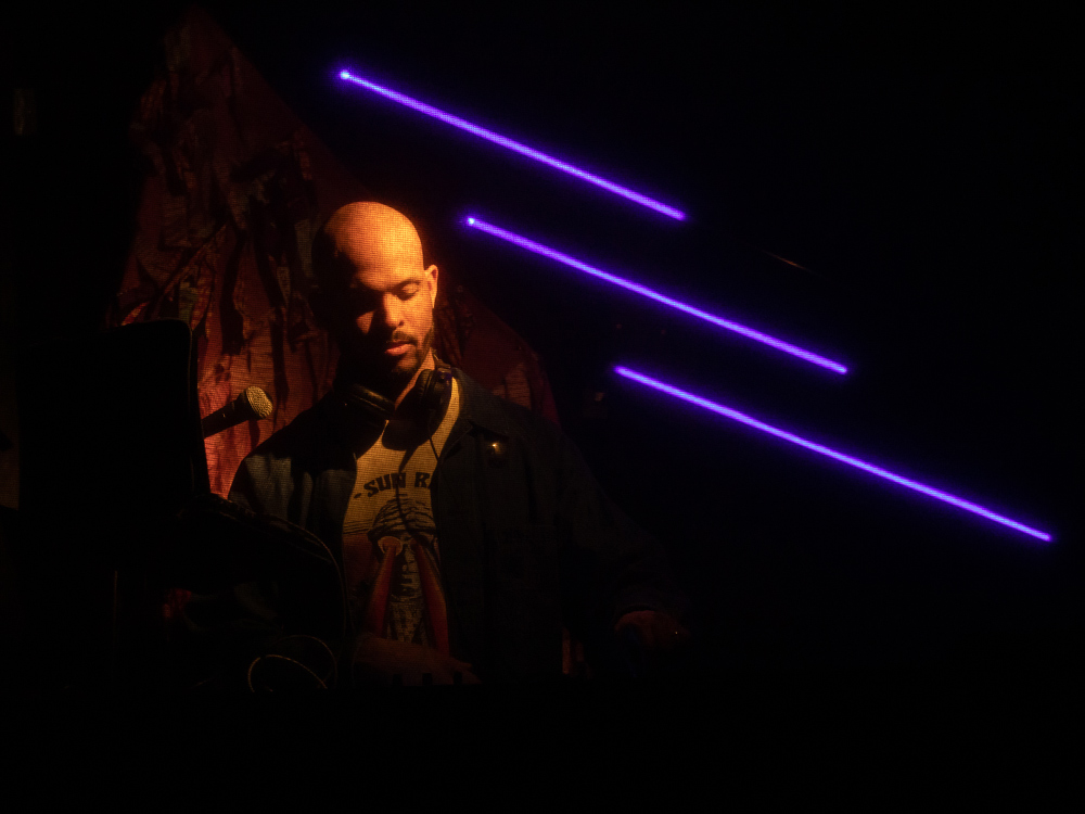 DJ Michael Manners DJs in the background, with purple neon light stripes behind him. He is wearing headphones around his neck.