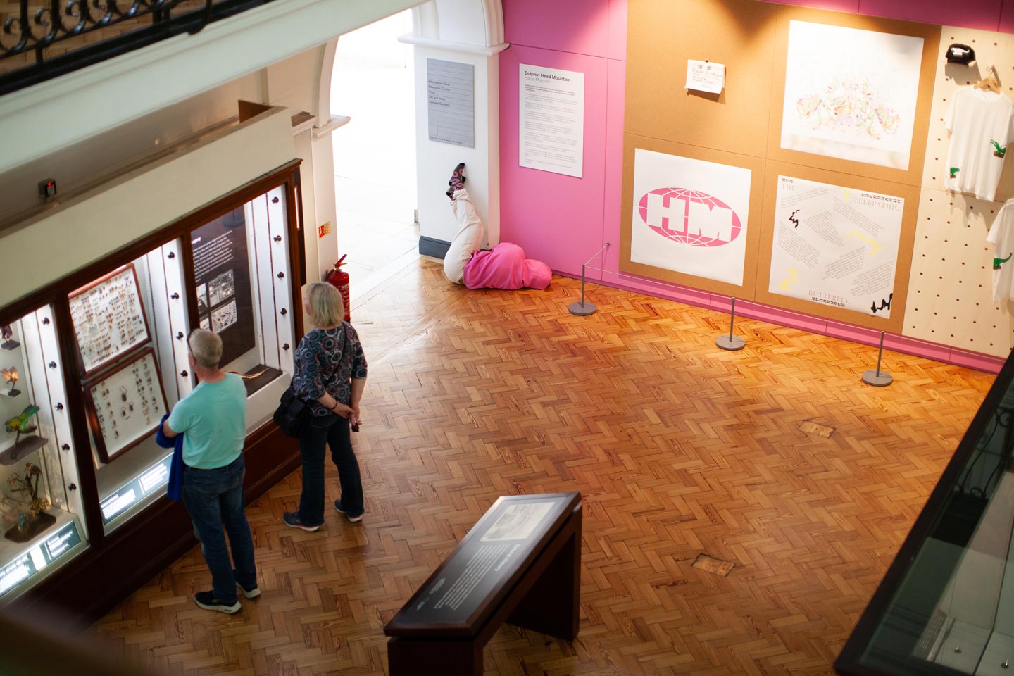 A shot of a gallery floor from a balcony. Two people look into a glass cabinet. In the background, a figure in pink and white clothes blends into the wall, against a pink and white branded display of logos and apparel.