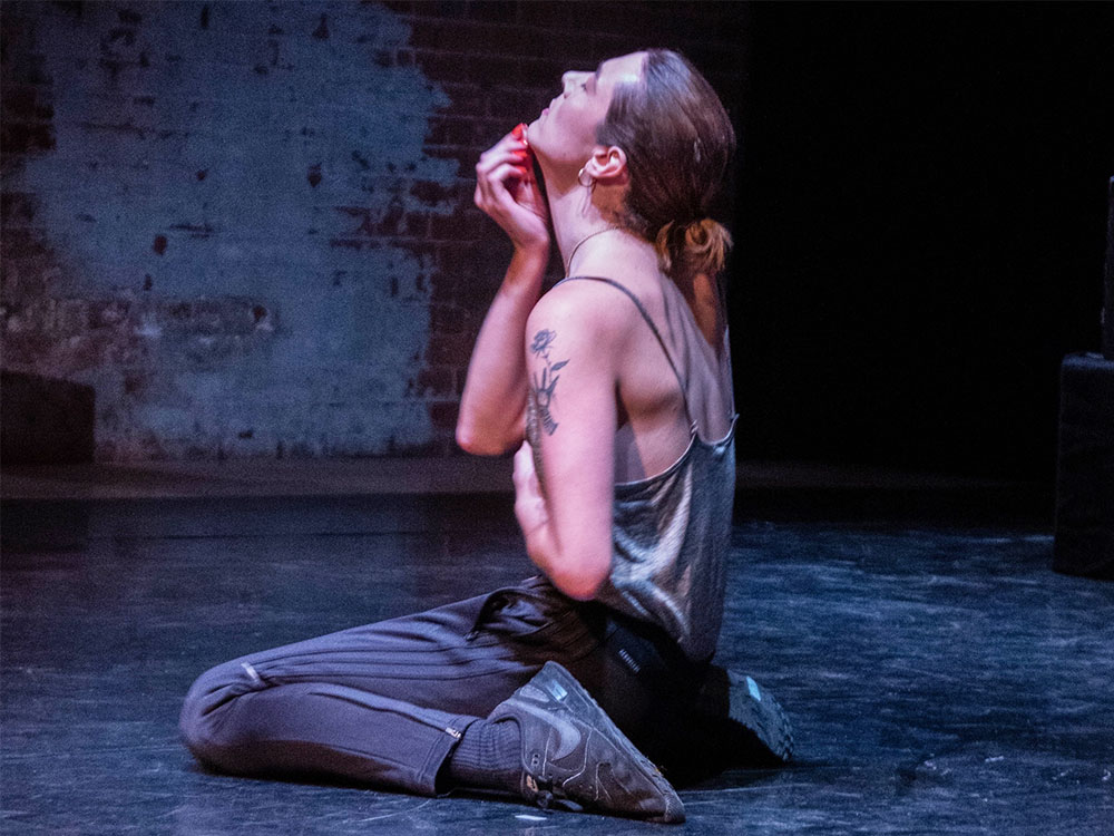 A white person in a singlet kneels on a floor, their arms posed, holding their chin