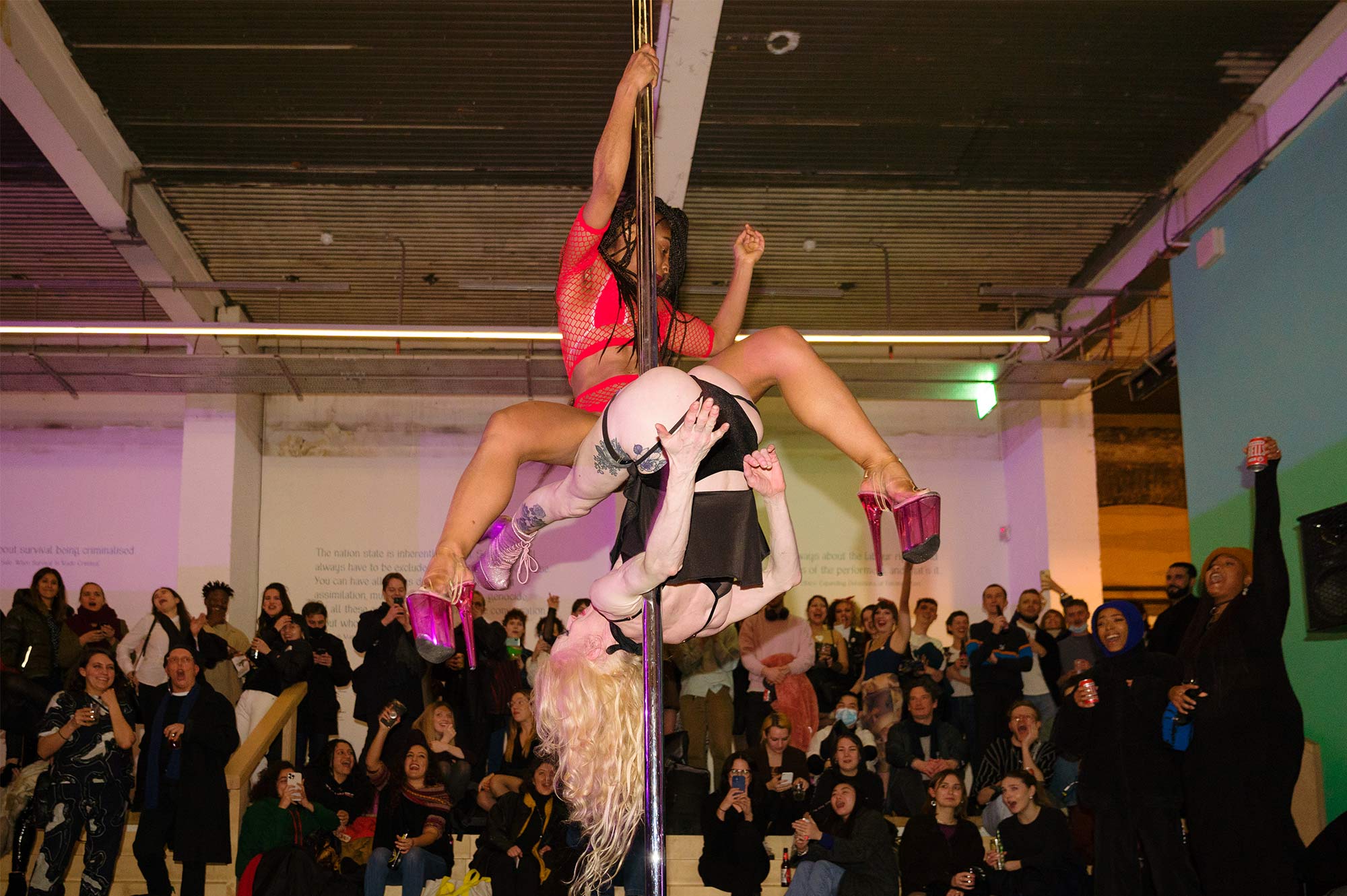 Two dancers in reverse, vertically, on a pole. A crowd cheers in the background.