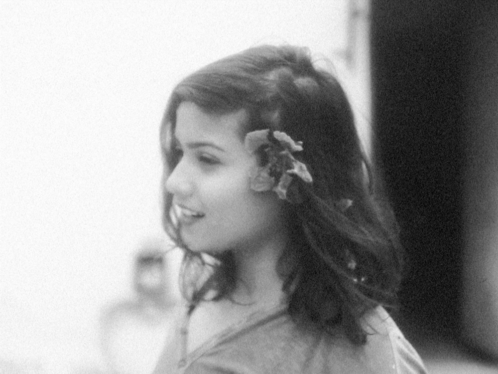 A black and white side portrait of a person with a flower in their hair, black-brown and flowing freely to their shoulders. They have an expression of joy.