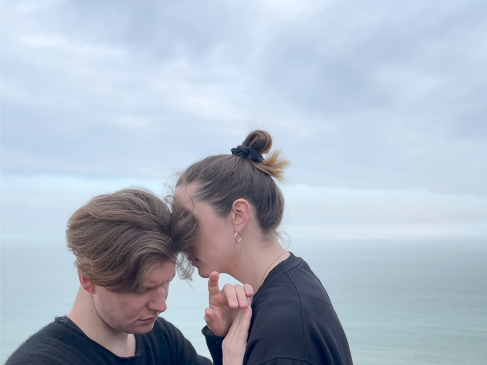 Two white people in embrace or dance against an ocean backdrop