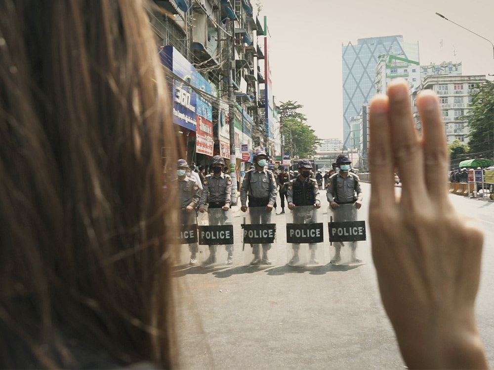 Woman holds three fingers up against riot police on empty street