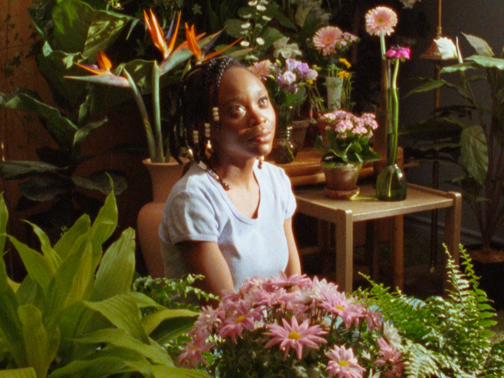 Black girl looks up longingly, in room surrounded by vibrant plants