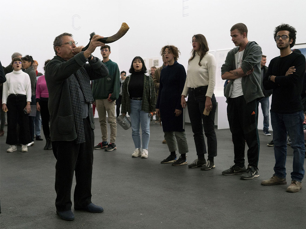 In a gallery space, a man blows into horn, surrounded by participants singing