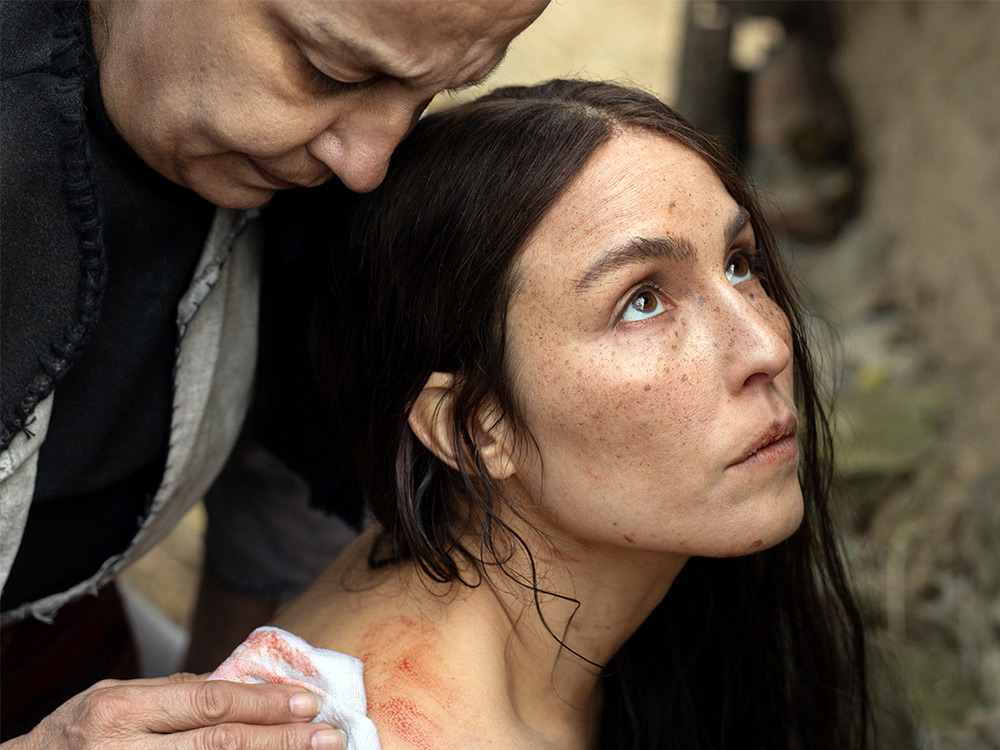 A person with long hair looks longingly to the sky. An older person wipes blood tenderly from the woman's shoulder.