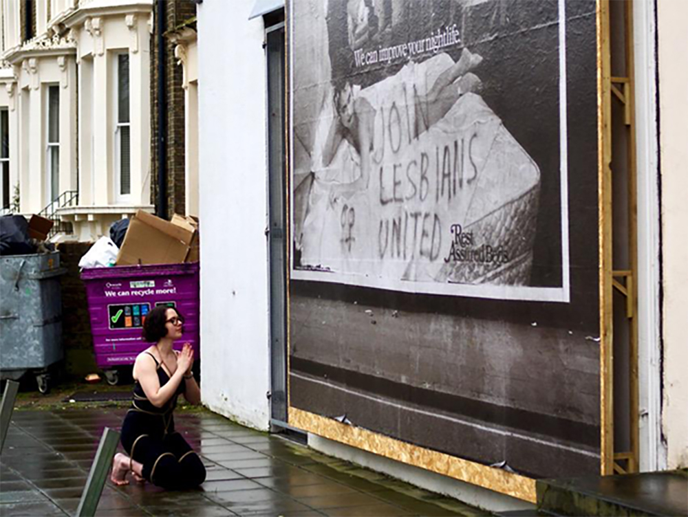 A person kneels on a wet London street in front of a billboard that reads 'JOIN LESBIANS UNITED'