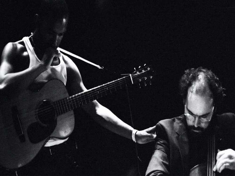 In greyscale, a musician in a singlet holds an acoustic guitar, in thought, while a cellist plays beside them.