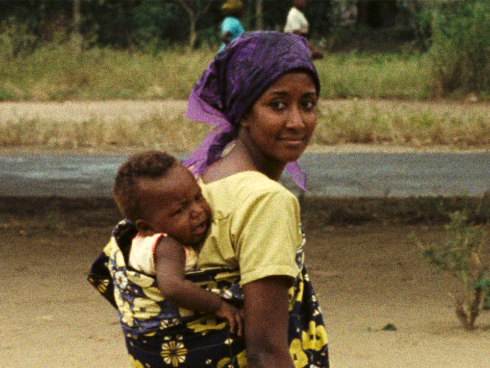 A mother carrying a child on her back, walking towards a road in a green and brown landscape
