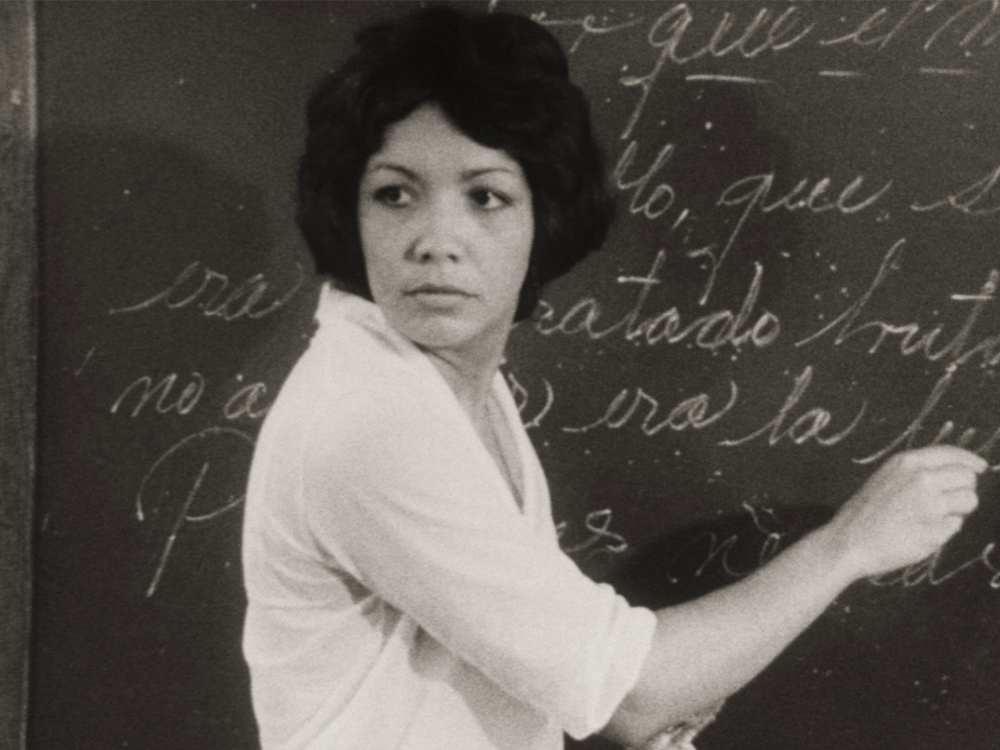 A woman turns back to look off-camera as she writes in Spanish on a blackboard