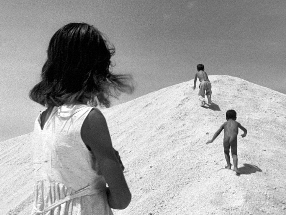 A person with shoulder-length hair stares off at two young kids running up a sandy hill