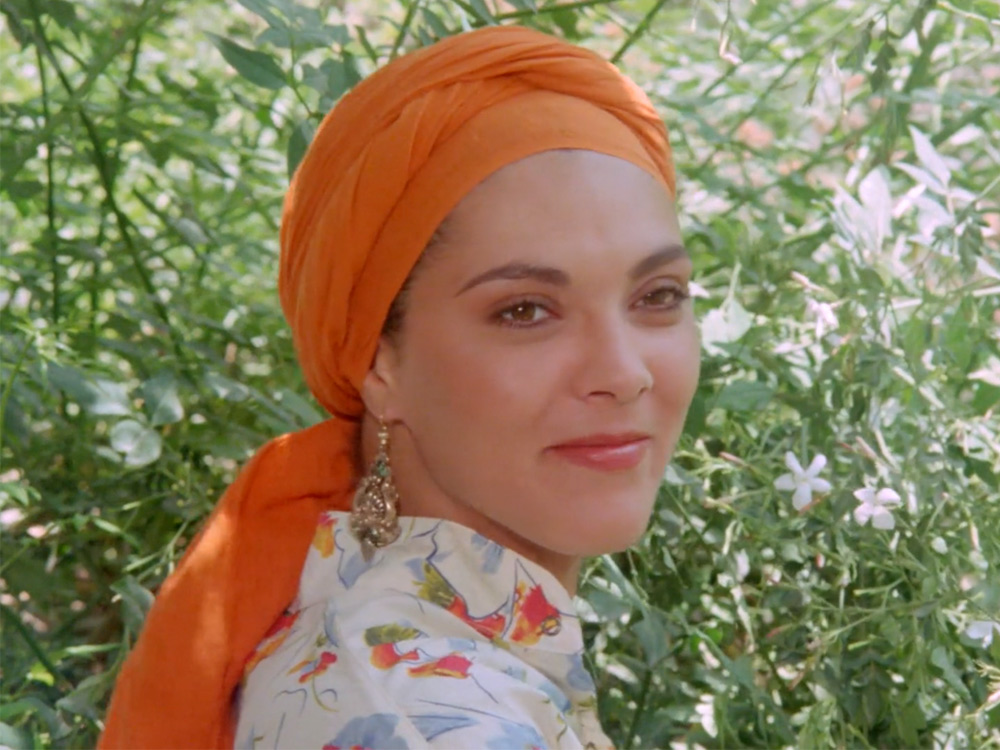 A person stares at the camera with quiet joy in front of bright green plants. She wears a bright orange headpiece