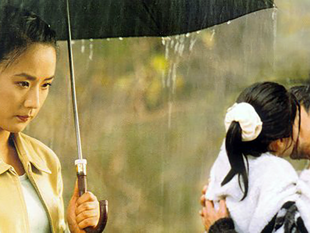 A woman stands under an umbrella in heavy rain, while beside her a couple embraces in a kiss