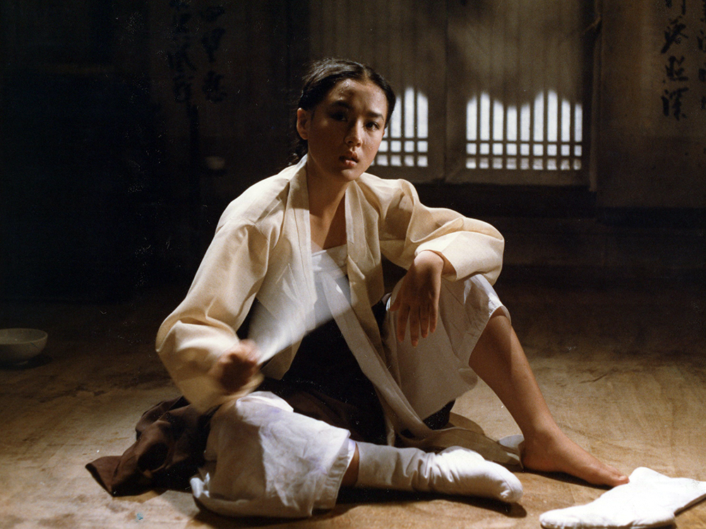 A Korean women in loose Joseon era clothing sits in a traditional room and stares towards the camera