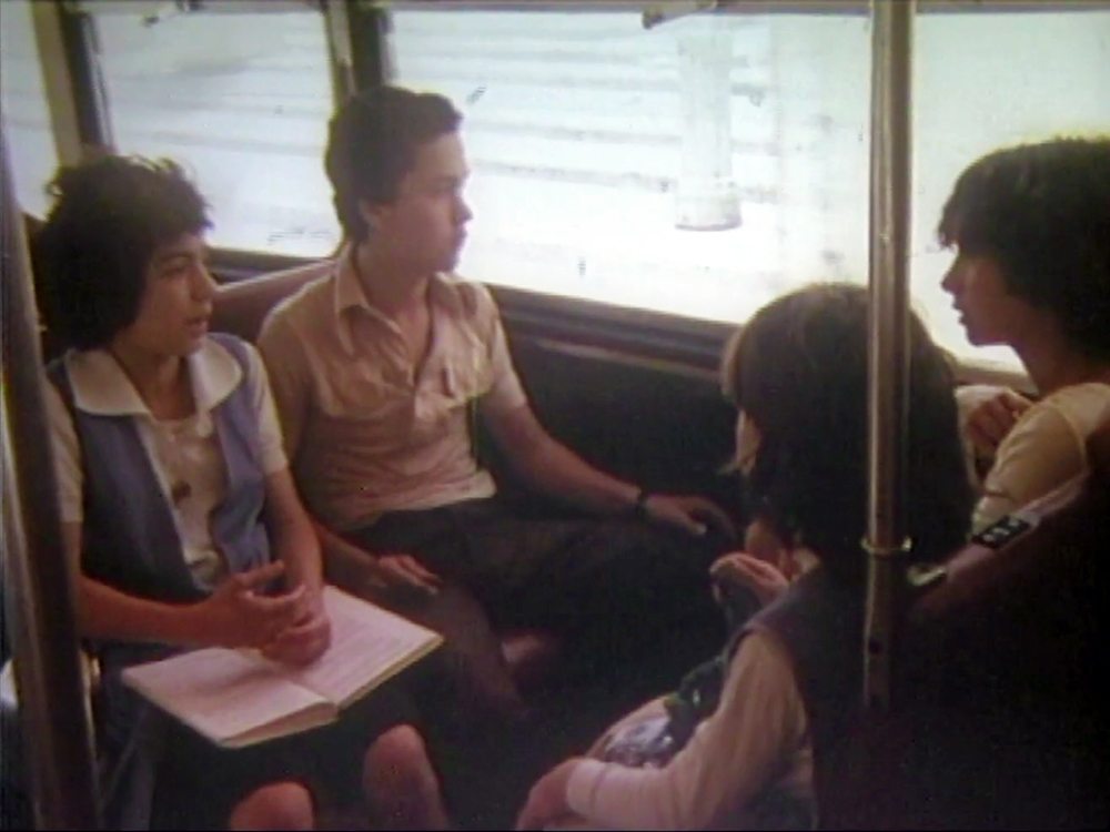 Four young people sit facing one another on a bus, on an old print of film