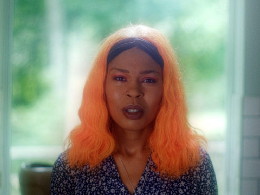 A young Black woman wears a headband and floral top, with neon orange hair. She stares straight into the camera, mouth slightly open, a look of stillness, but with direction