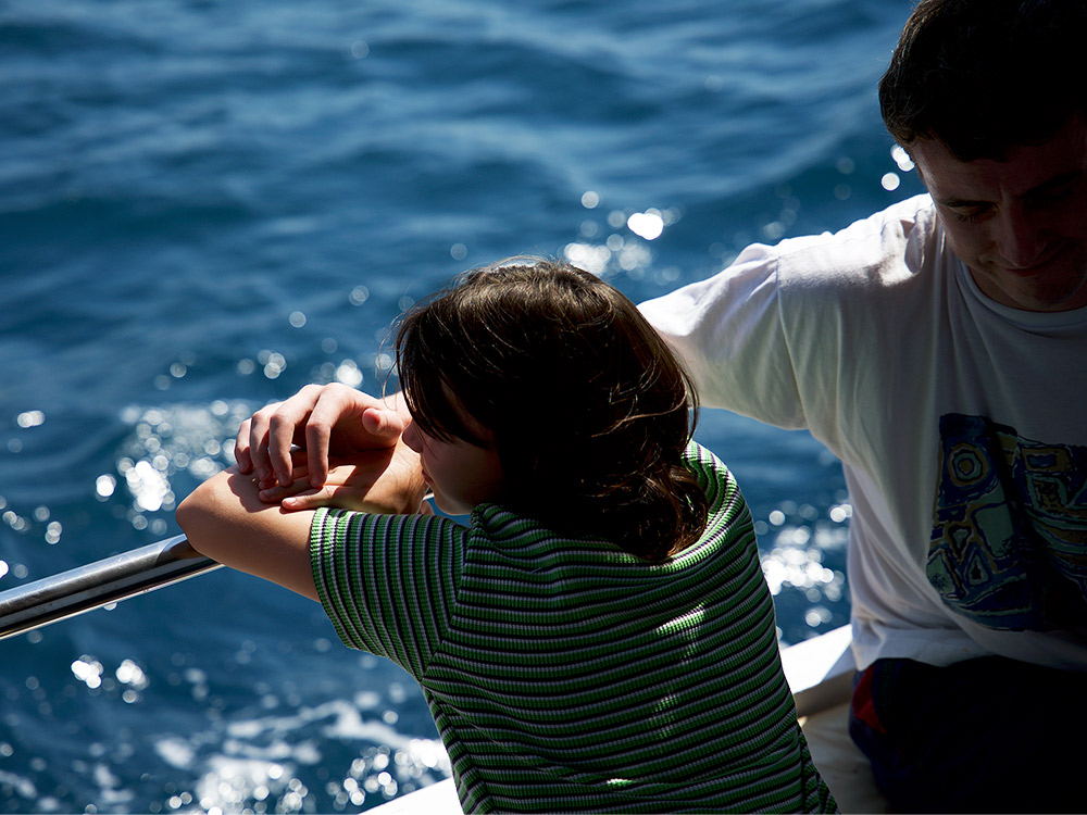 A young couple sit by the edge of a boat, a young person staring off into the distance while their friend/partner holds a tender arm across