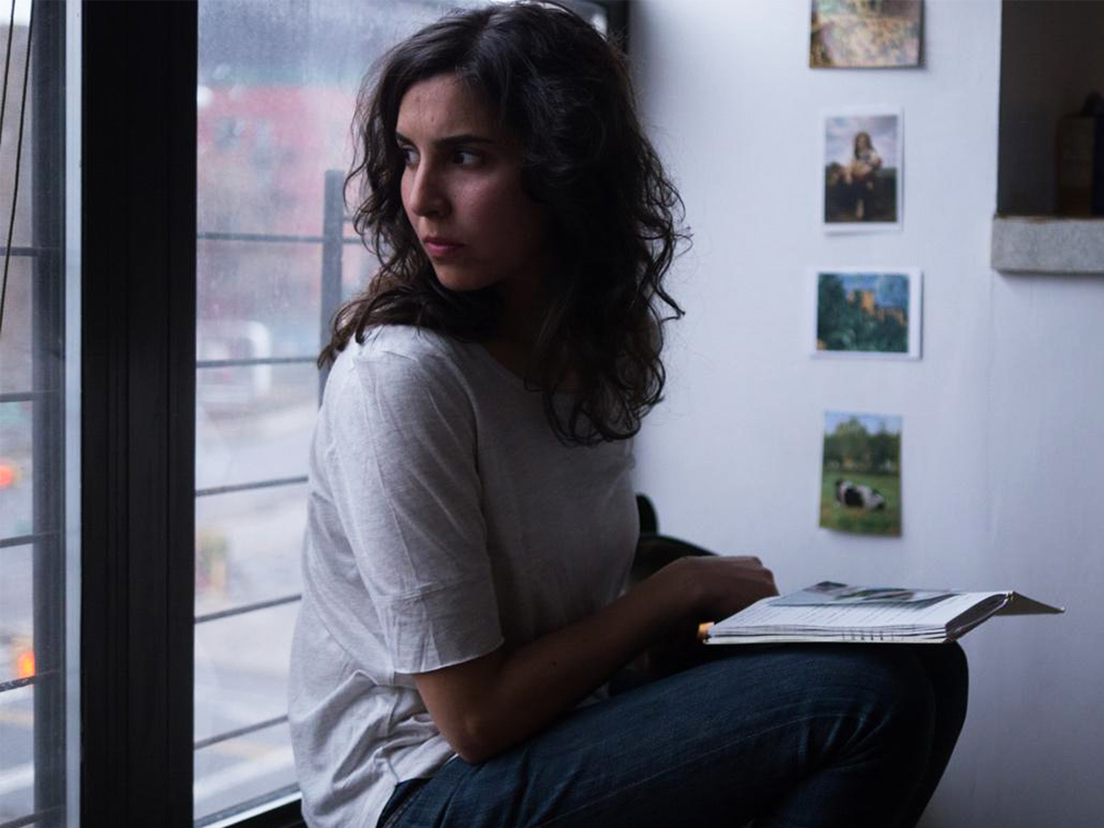 A woman sits by the window, looking out to the street. Behind her are photographs. She has a notebook and a photograph on her lap.