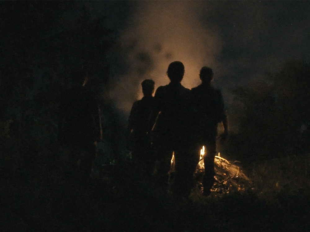 Four figures stand in front of a burning fire outside at night.