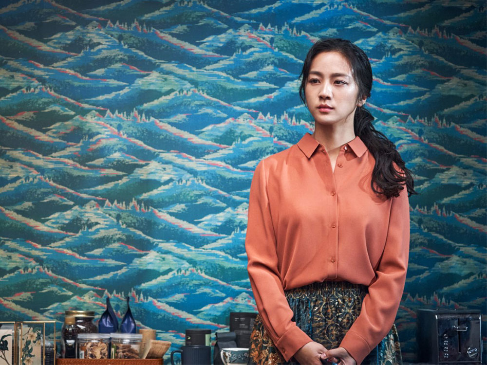 A woman in a salmon-red top stands against a wallpaper of mountains. Behind her are well-curated objects: tea, coffee cups. She is looking still, like she is keeping secrets to herself.
