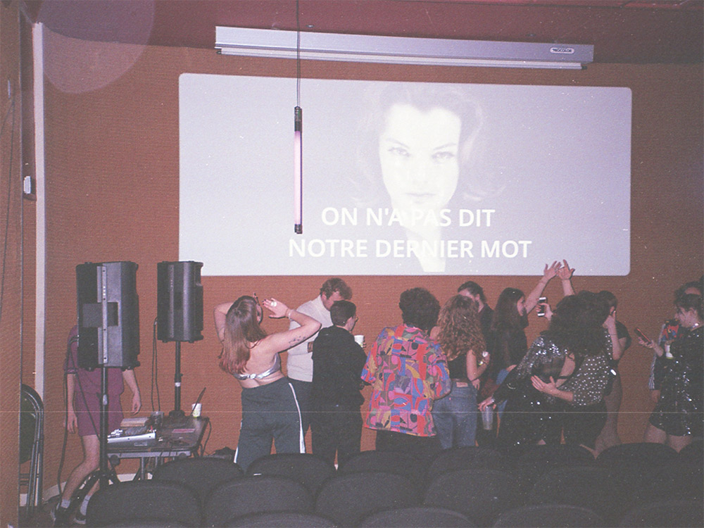 A group of young people dance in front of a cinema screen while a musician plays a set to the side