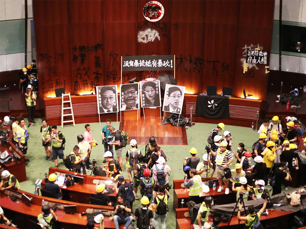The Hong Kong Legislative Council building is sprayed with black Chinese writing and portraits of members of the Council. An assembly of press in hardhats record the spectacle.