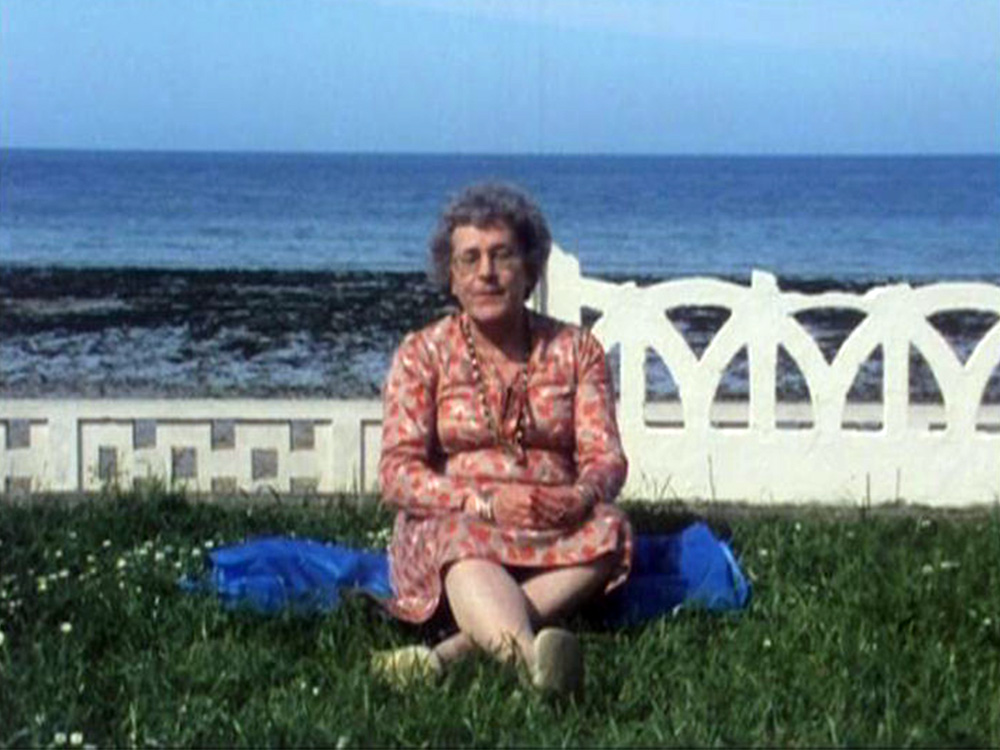 An elderly woman sits peacefully on a patch of grass by the ocean
