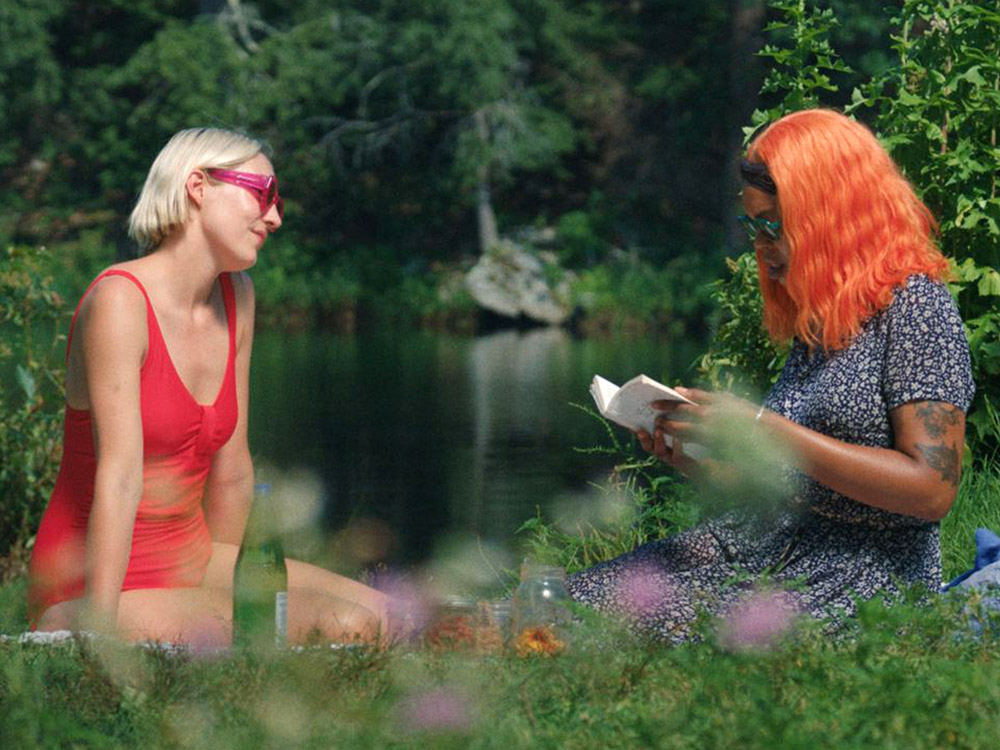 A person in a red swimsuit sits with her friend by the lake, reading a book together