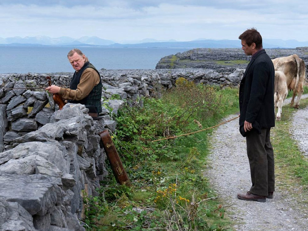 Two men stand at a rocky wall by a cliff. The distance is foggy. The older man, working at the wall, looks hostile
