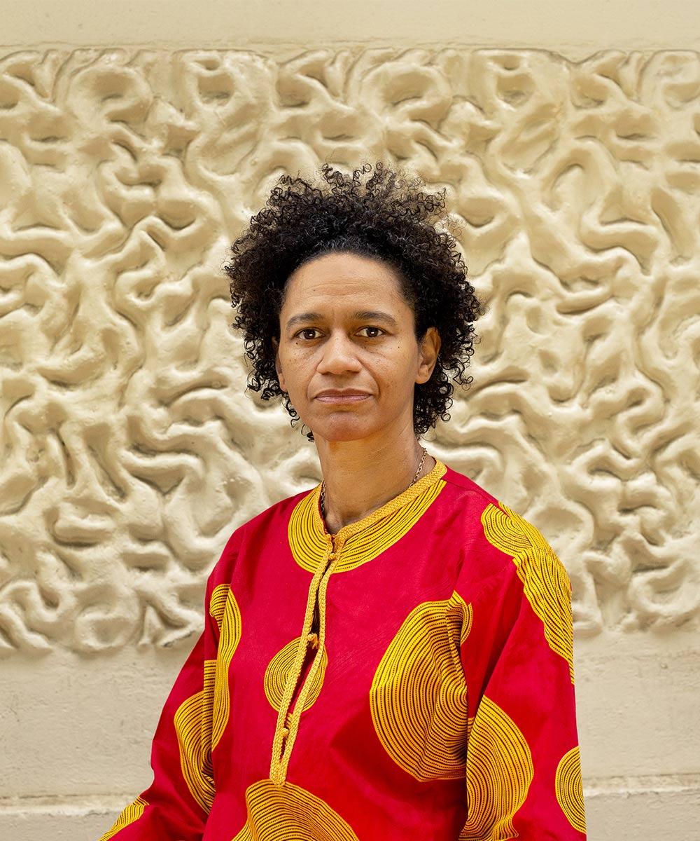 An image of Mojisola Adebayo. Photo taken by Bettina Adela. Mojisola is a black woman wearing a bright red and yellow jumper, sitting against the architectural features of the ICA outside walls