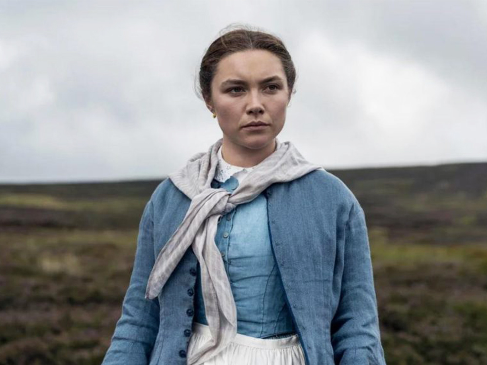 A woman stares into the distance, serious and contemplating. Behind her is a gothic grey sky and rugged landscape