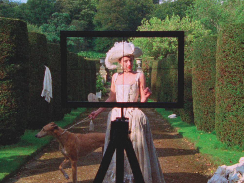 A person in a white dress stands in front of well-trimmed hedges. They have a long dog on a leash. A sight, like a camera or gun sight, is aiming at them.