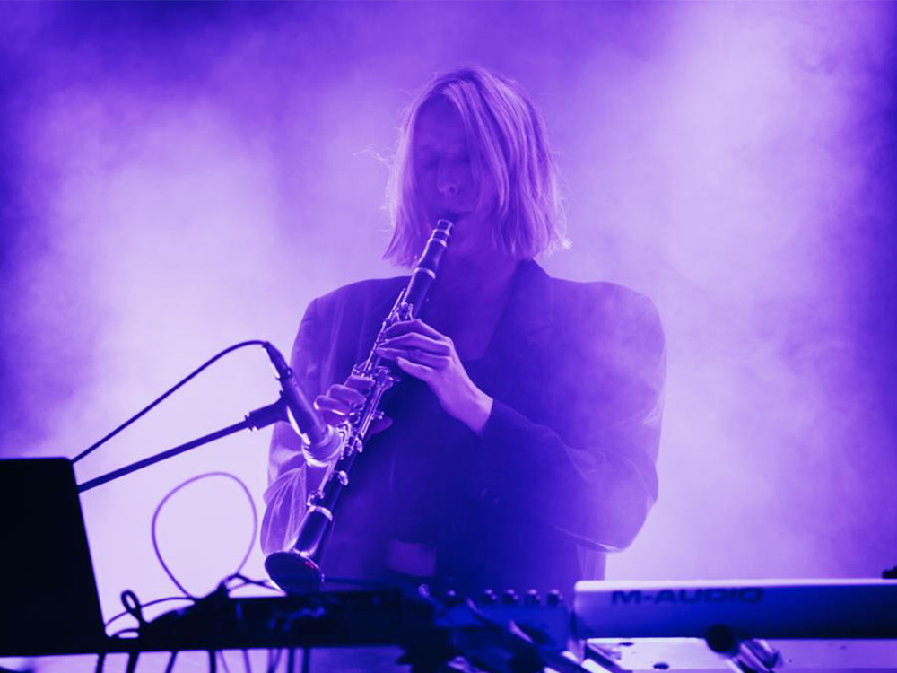A person with short hair plays a clarinet into a mic, in front of synths and a laptop. They are surrounded by dark blue fog
