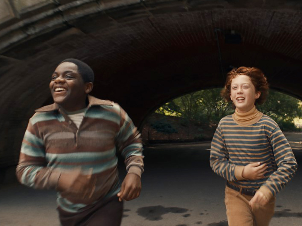 Two young kids, a black kid and a white kid, run with joy through a tunnel. They're wearing American 80s style clothes