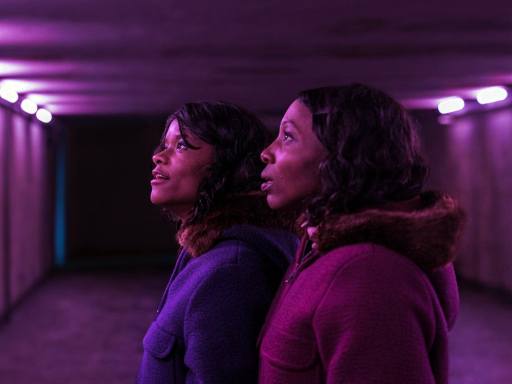 Two Black girls look off-camera in a tunnel bathed in purple light