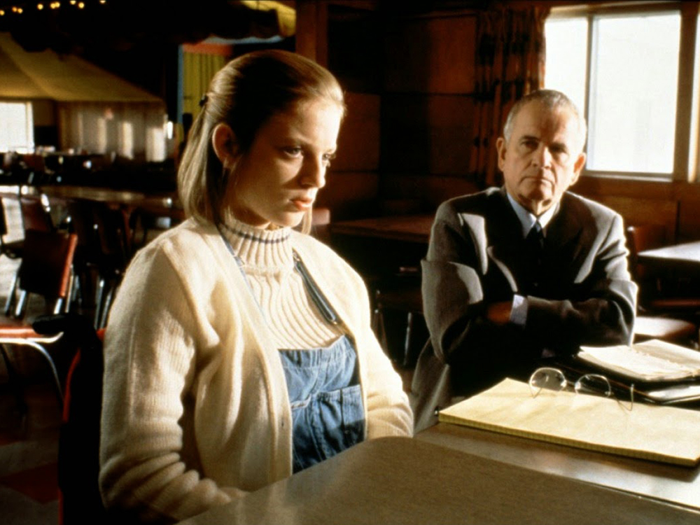 A young girl sits at a table looking sad, while an older man watches on