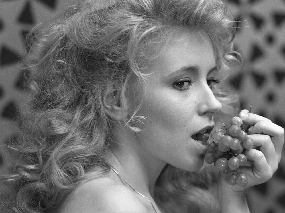 A black and white image of a feminine person with curled blonde hair, eating grapes and staring off-screen