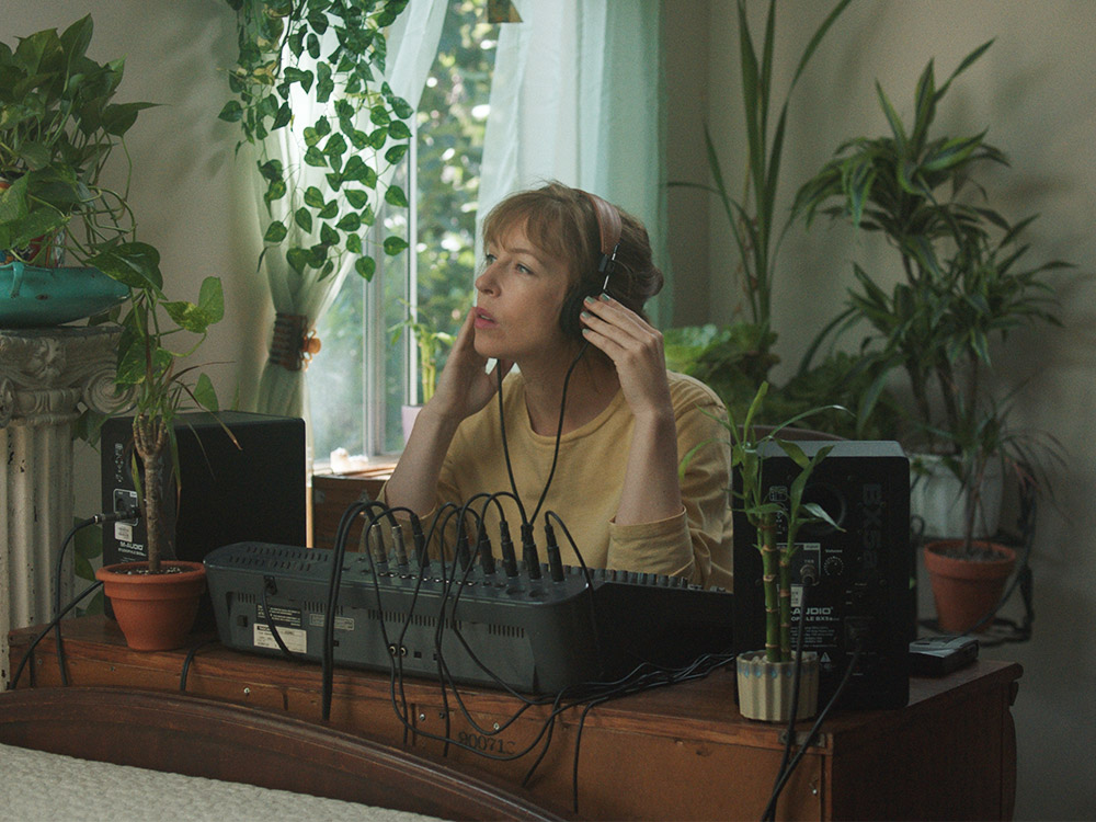 A person wears headphones, listening deeply to recordings coming out of a mixer. They are surrounded by house plants in a well-lit room