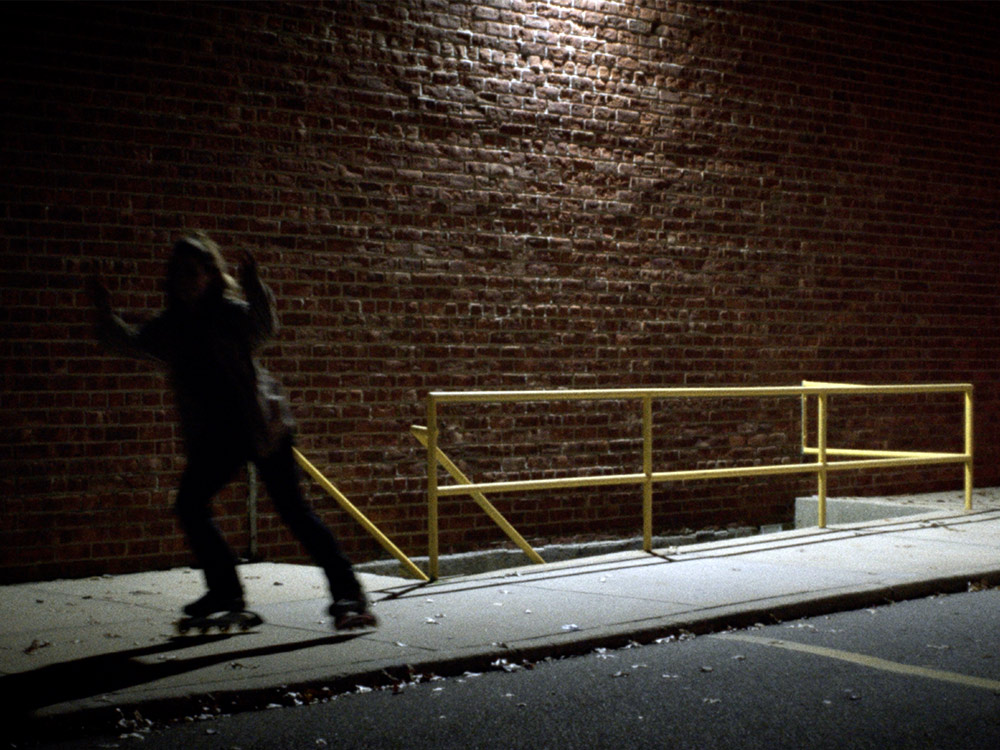 A person in shadows on rollerskates skates along a footpath, past a brick wall lit by a single light