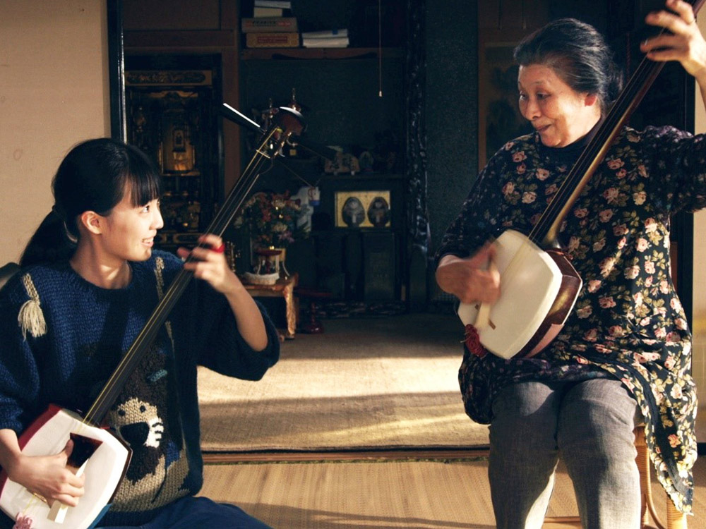A younger and older person play shamisen together joyfully in a traditional Japanese house setting