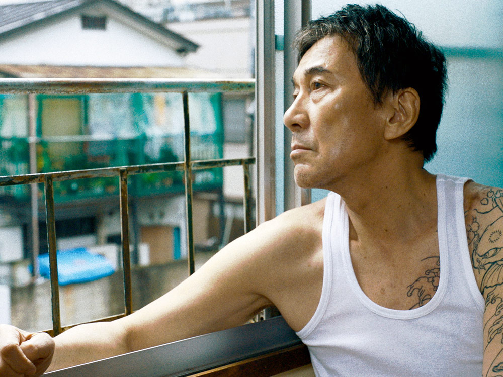 A tattooed person in a singlet stares longingly out a barred window