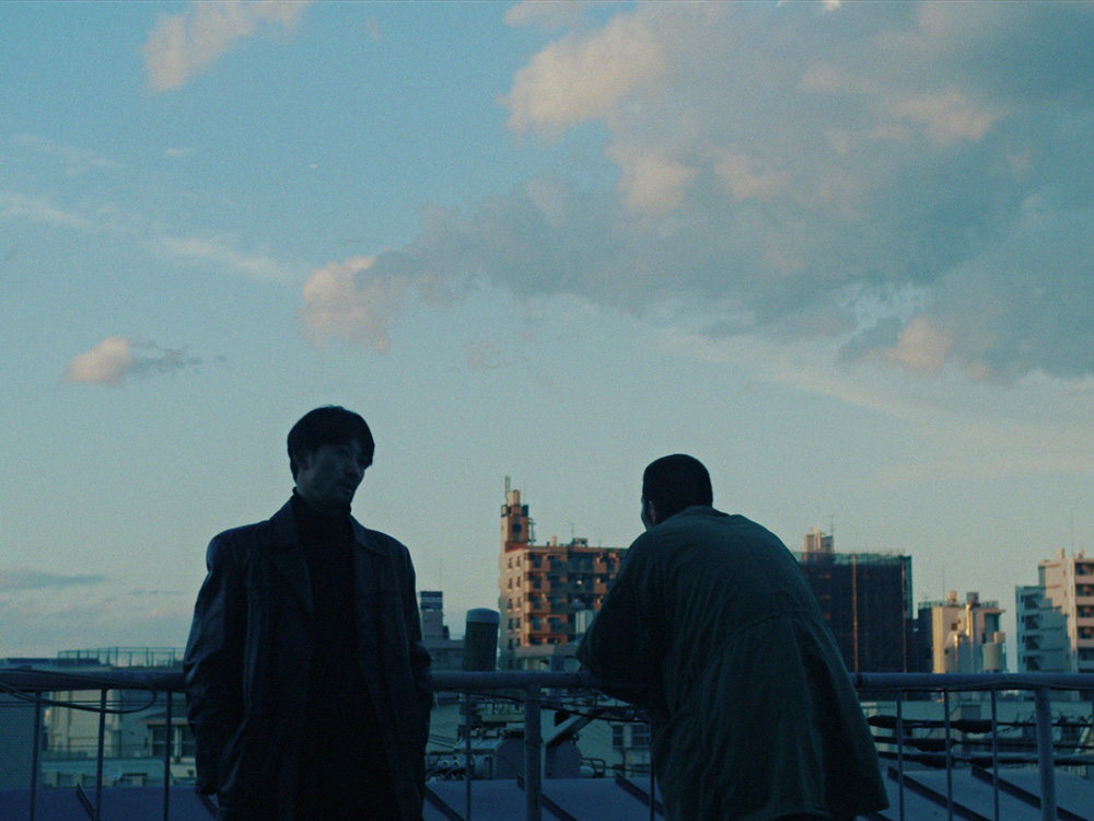 Two shadowy people in overcoats stand on a rooftop and stare out into a clouded sky