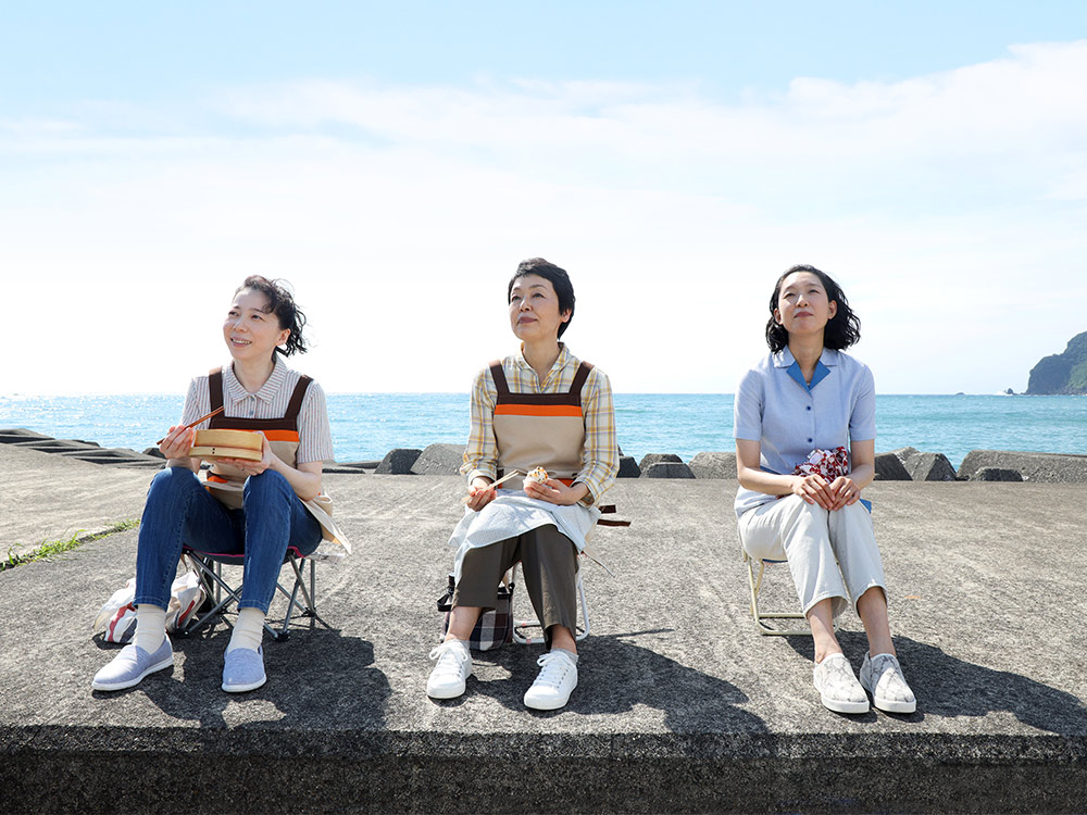 Three Japanese people wearing aprons stare out into the sky, eating lunch, the ocean behind them