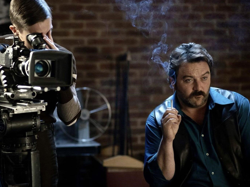 A man smokes and watches his movie set, expressionless. The cameraman films from the side, poised