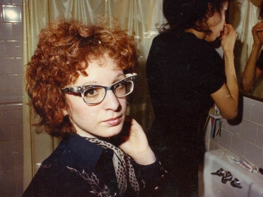 A grainy photo of Nan Goldin. She's got bright red hair, glasses, staring straight into the camera with her hand beside her chin. In the background, someone - maybe her flatmate - is doing their morning/evening bathroom rituals