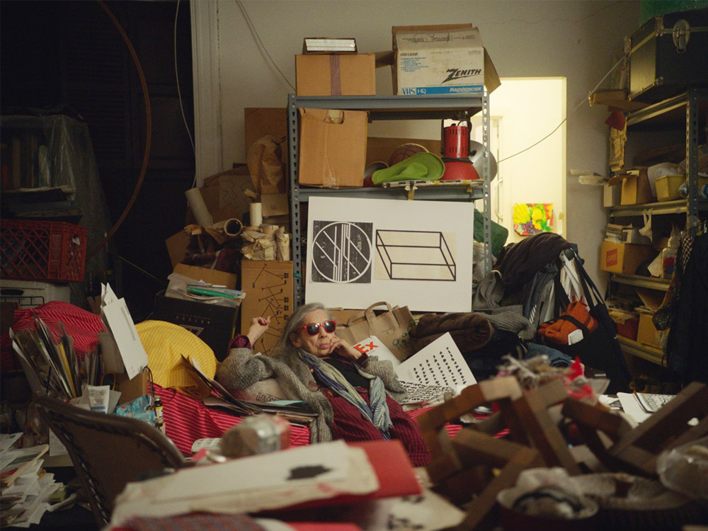 A person with grey hair in a fur coat and red aviators sits in the centre of piles of objects, books, paintbrushes, boxes and shelves in their apartment