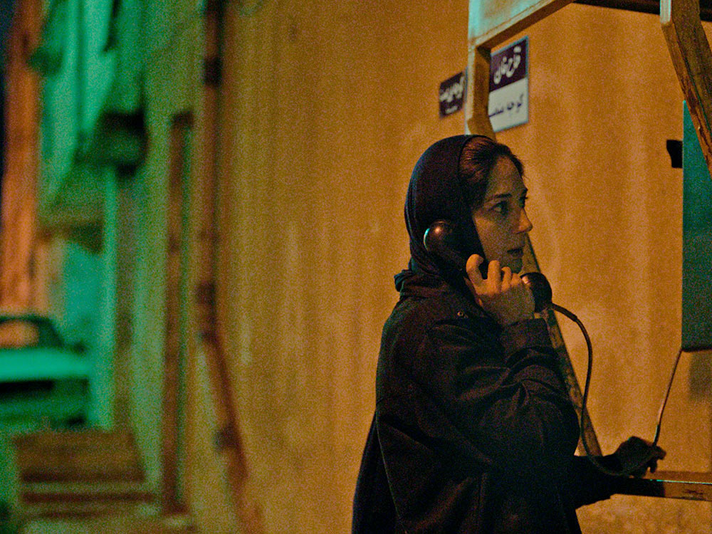A person takes a call from a public telephone in a quiet street of Mashhad. They have a look of slight surprise on their face.