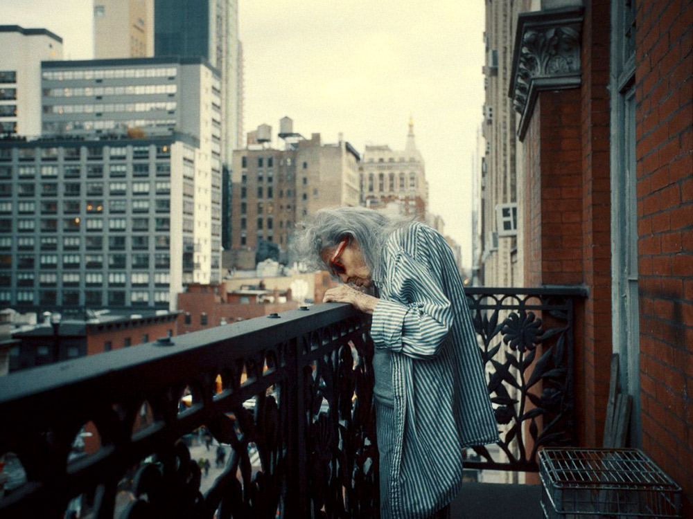 An elderly resident of the Chelsea leans over the balcony of their hotel in the Chelsea Hotel, backdropped by tall New York buildings